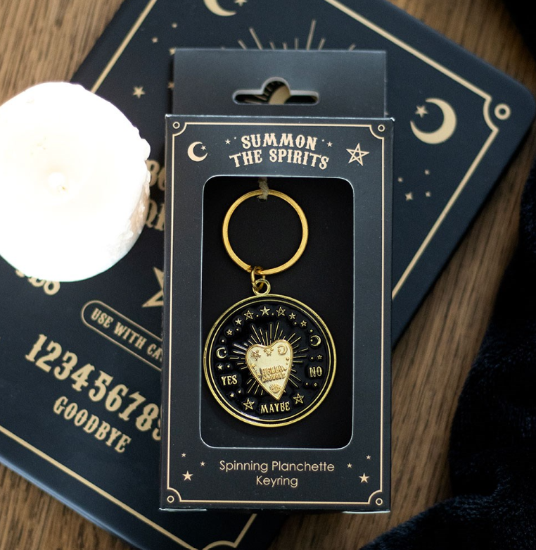 A gold-toned spinning planchette keying charm in its black retail packaging, displayed on a dark celestial-themed card over a wooden surface with a white candle partially visible.