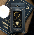 A gold-toned spinning planchette keying charm in its black retail packaging, displayed on a dark celestial-themed card over a wooden surface with a white candle partially visible.